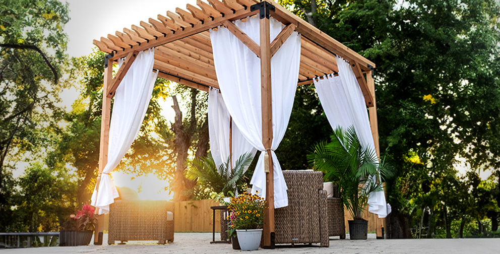 Outdoor wooden pergola with white drapes, surrounded by plants and wicker furniture, set against green trees and sunlight in the background.