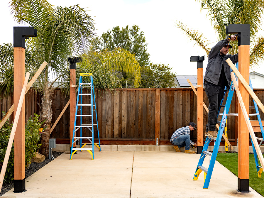 Workers installing a wooden pergola structure using Simpson Strong-Tie Outdoor Accents hardware, with ladders and bracing in a backyard setting.