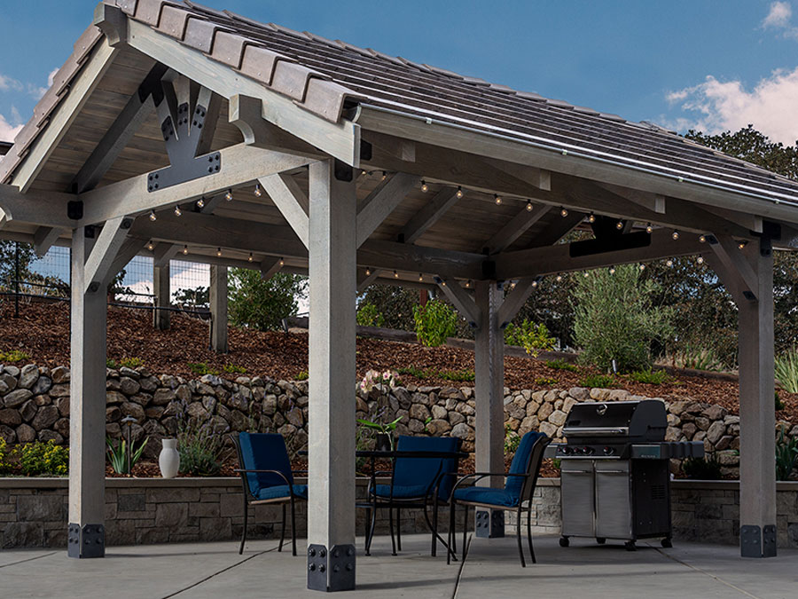 Outdoor pergola with wooden beams and metal connectors, set up on a patio with a barbecue grill and seating area.
