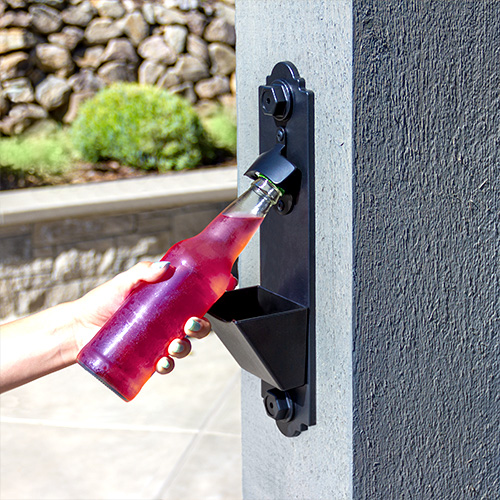 Hand opening a pink bottled drink with a mounted bottle opener and cap catcher on an outdoor post.