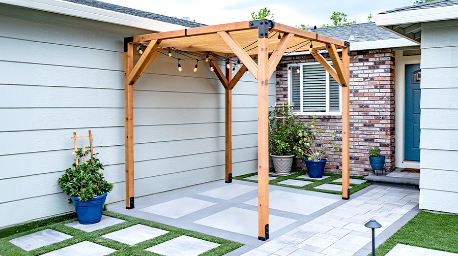 A wooden wall-mounted pergola with black metal brackets, string lights, and potted plants in a neatly landscaped backyard.