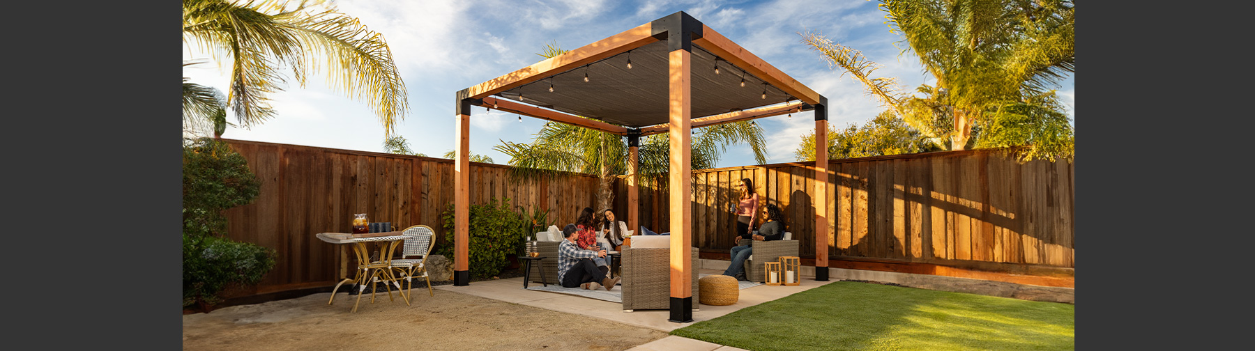 A modern pergola with black metal connectors and cushioned seating, set on a concrete patio surrounded by greenery.