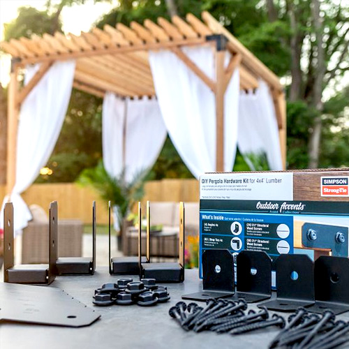 Pergola hardware kit with brackets, fasteners, and packaging on a table, with a wooden pergola and white drapes in the background.