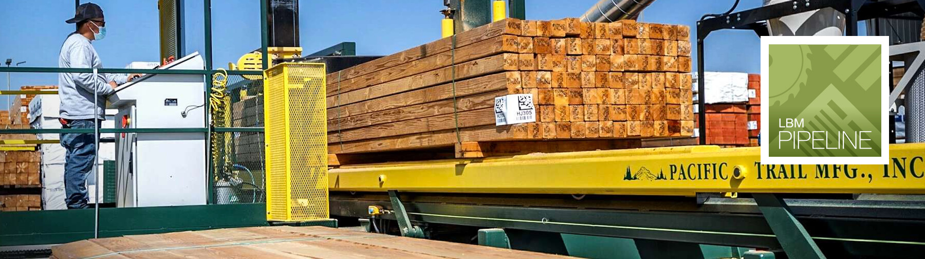 Worker operating control equipment near a large stack of bundled lumber on a yellow machine platform outdoors.