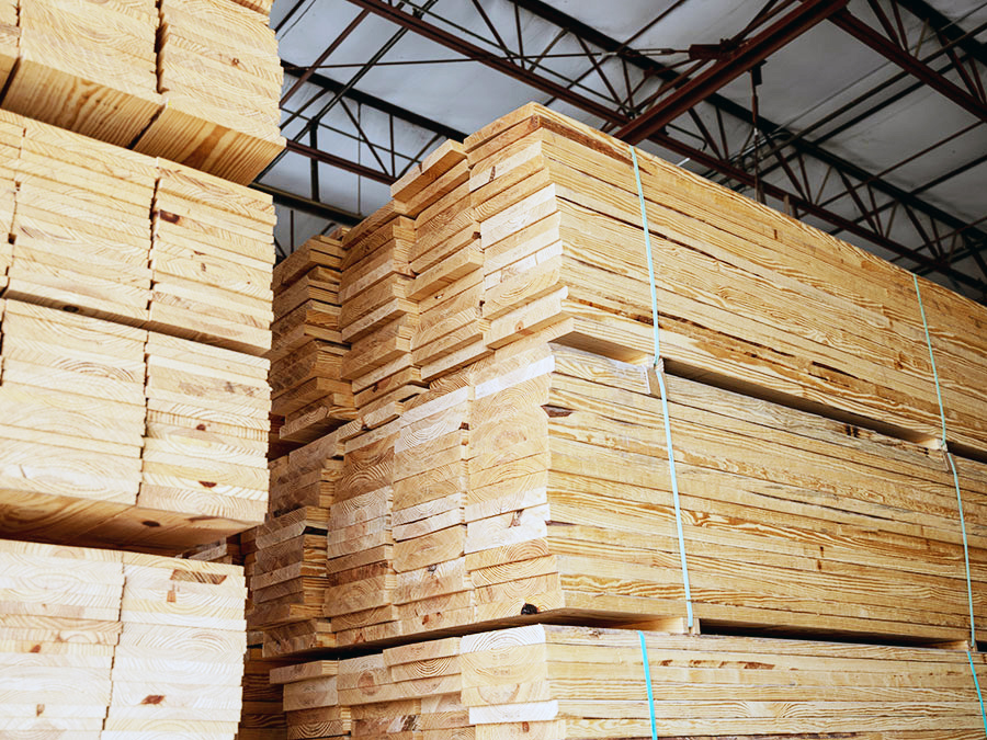 Stacks of neatly bundled lumber stored in a spacious warehouse with metal roof beams.
