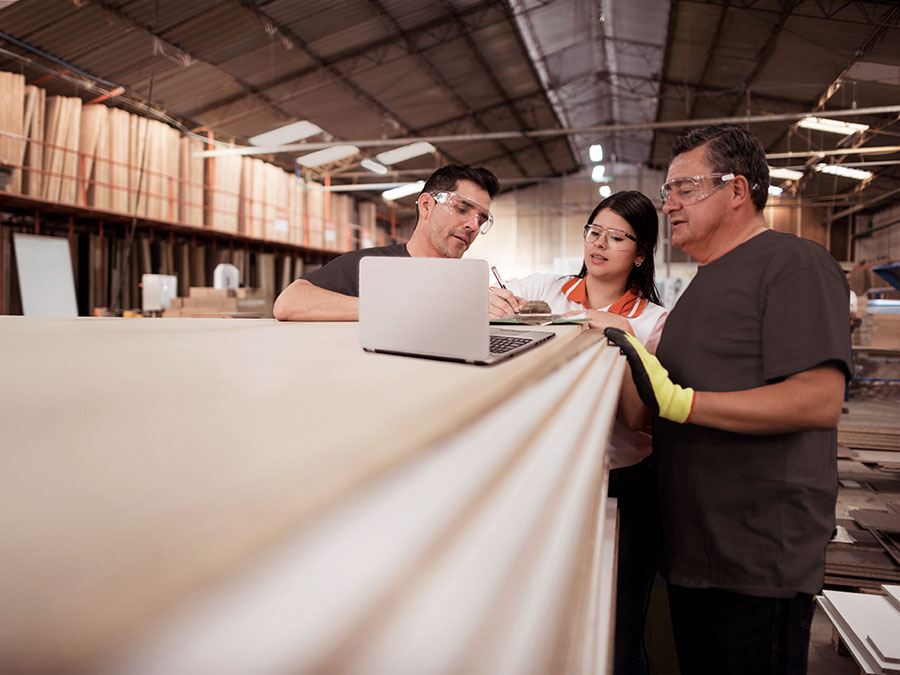 Three workers in safety glasses collaborating in a wood manufacturing warehouse, reviewing data on a laptop.