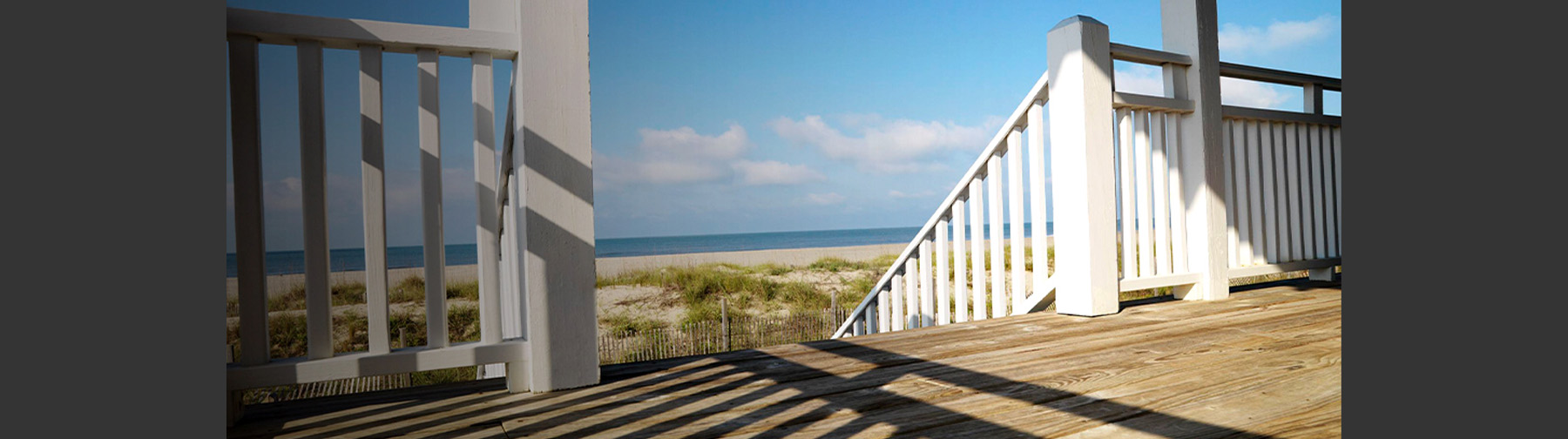A beachside wooden deck with white railings and posts overlooks sand dunes and the ocean under a bright blue sky.