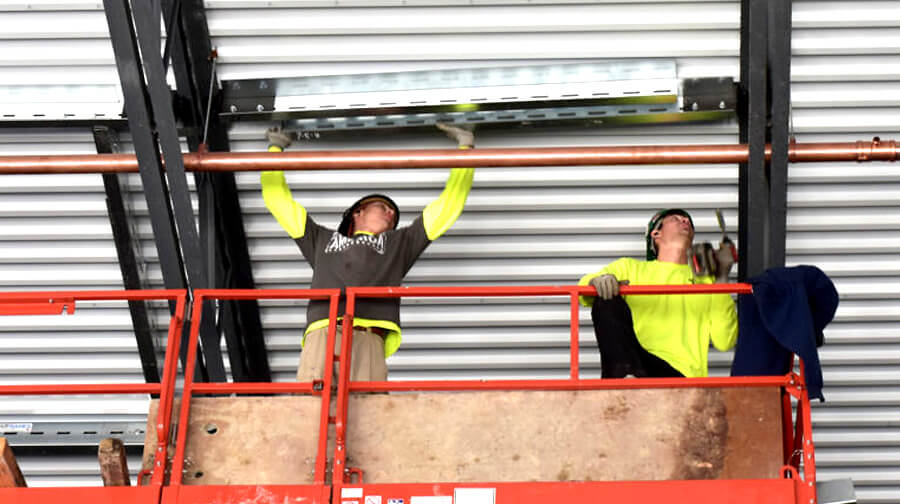 Two construction workers on an elevated lift install a metal frame on the ceiling, securing it to the steel structure of a building.