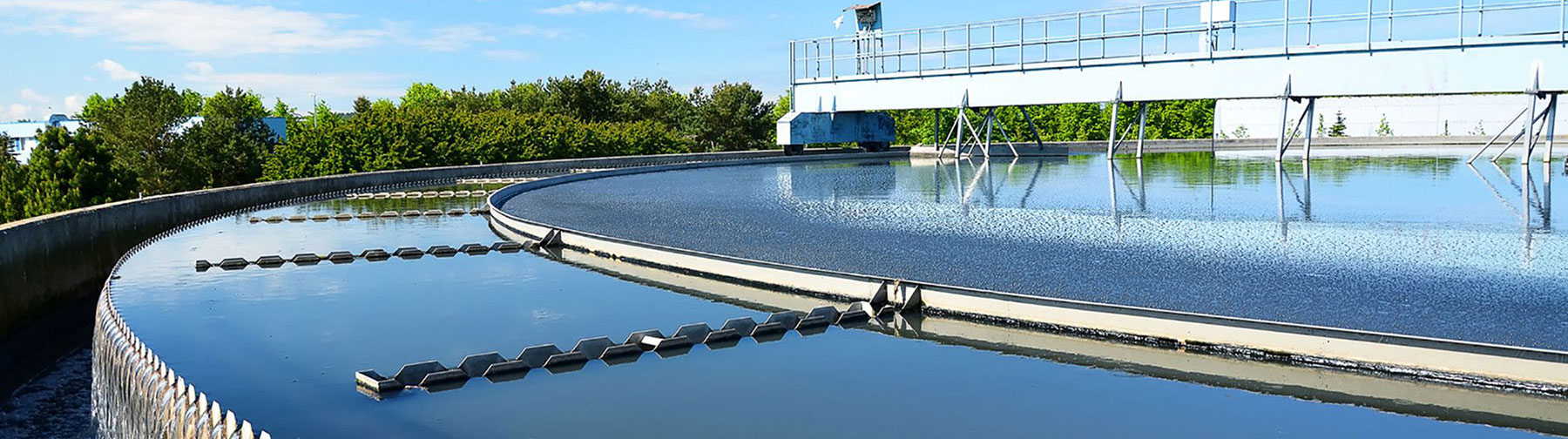 Overview of a large circular water treatment structure at a wastewater facility, surrounded by trees on a clear day.