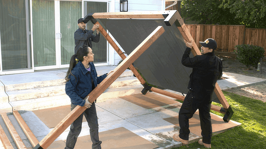 Three people lifting and assembling a pergola frame in a backyard, aligning wooden beams and metal connectors on a concrete patio.
