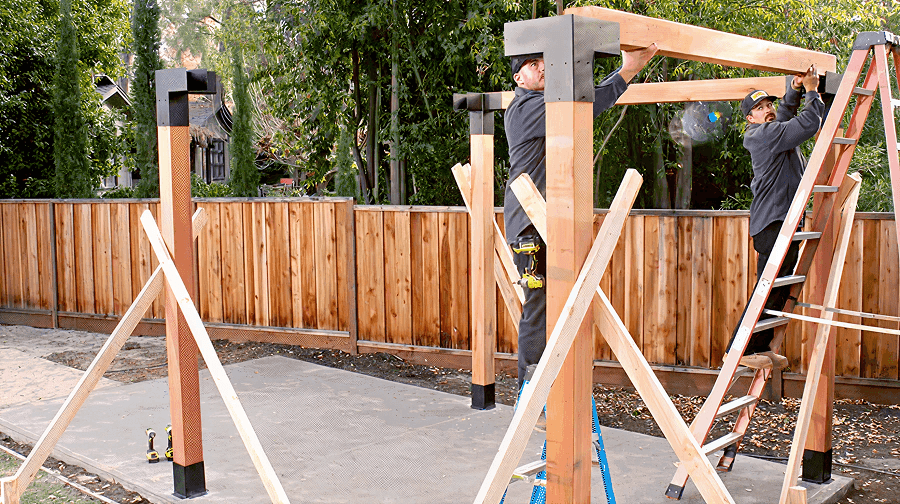 Two builders on ladders installing beams on a pergola structure, supported by braced posts on a concrete slab.