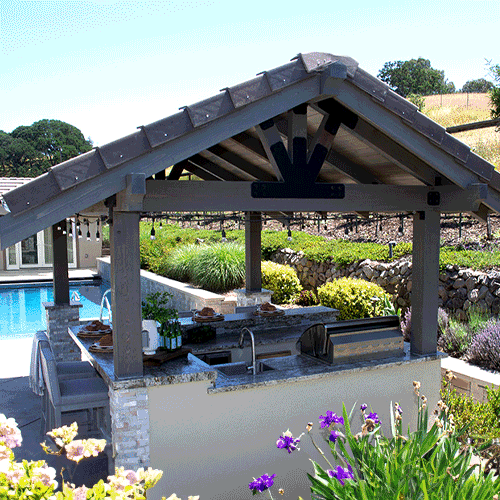 An outdoor kitchen under a wooden pavilion with black metal accents, featuring a grill, countertop, and a scenic garden view.