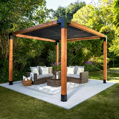 A modern pergola with a wooden frame, black metal accents, and a shaded top shelters a cozy wicker seating area in a lush green backyard.