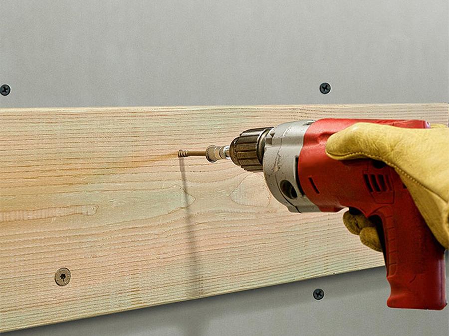 yellow gloved hand using red handrill to drill SDWS Timber screw into wood slab and grey wall