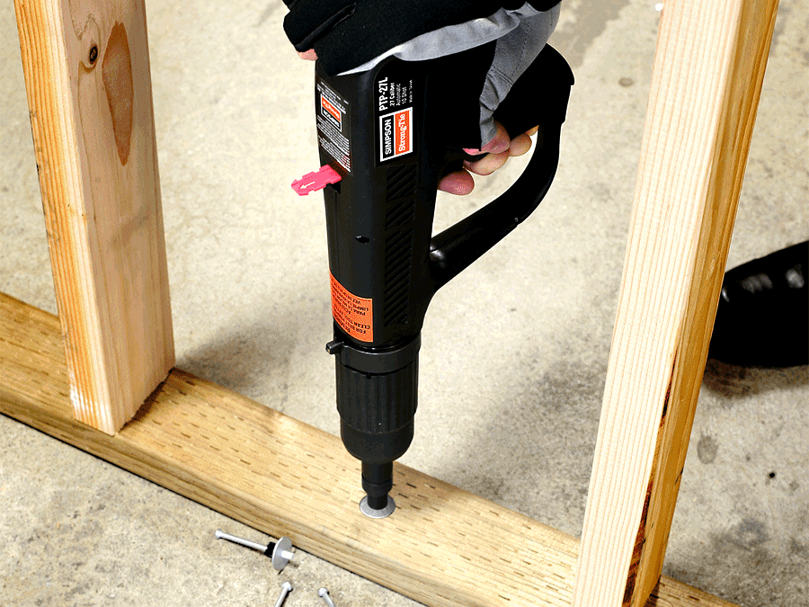 Close-up of a Simpson Strong-Tie PTP-27L powder-actuated tool being used to drive a fastener through a wood plate into concrete, shown in a certification or training setting.