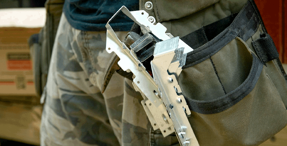 Close-up of a construction worker’s tool belt holding a Simpson Strong-Tie metal connector, with packaging and materials visible in the background.