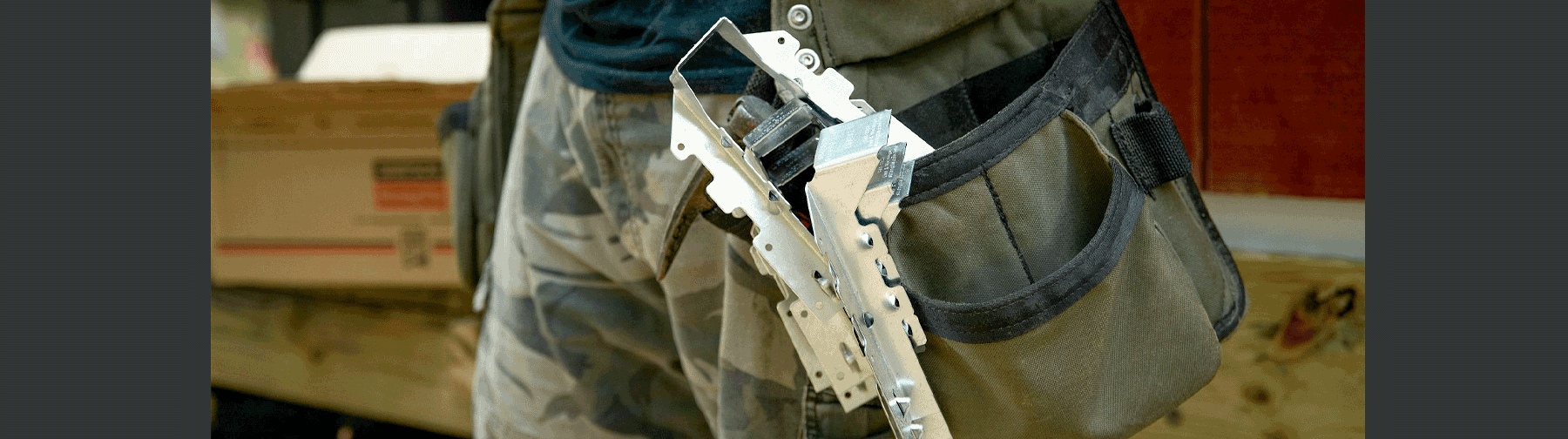 Close-up of a construction worker’s tool belt holding a Simpson Strong-Tie metal connector, with packaging and materials visible in the background.