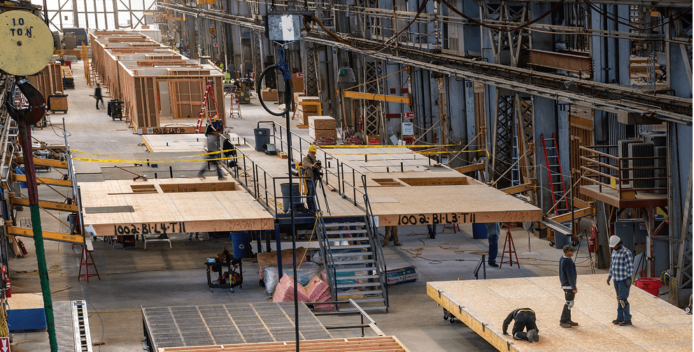 Indoor construction site with wooden frames, workers, tools, ladders, scaffolding, and yellow caution tape scattered throughout.