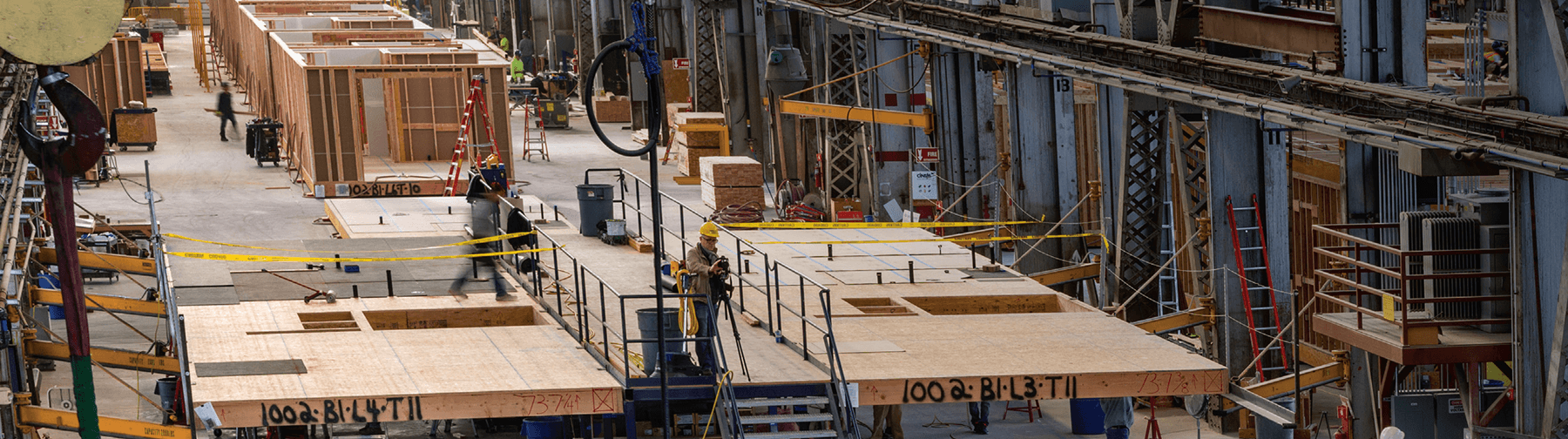 Indoor construction site with wooden frames, workers, tools, ladders, scaffolding, and yellow caution tape scattered throughout.