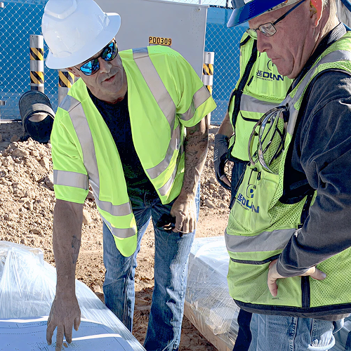 Construction workers wearing safety vests and hard hats review blueprints at a job site, discussing project details near wrapped building materials.